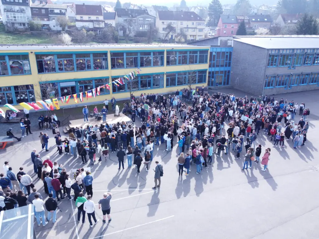 Luftaufnahme: Sehr viele Menschen versammeln sich auf dem Schulhof vor dem Schulgebäude