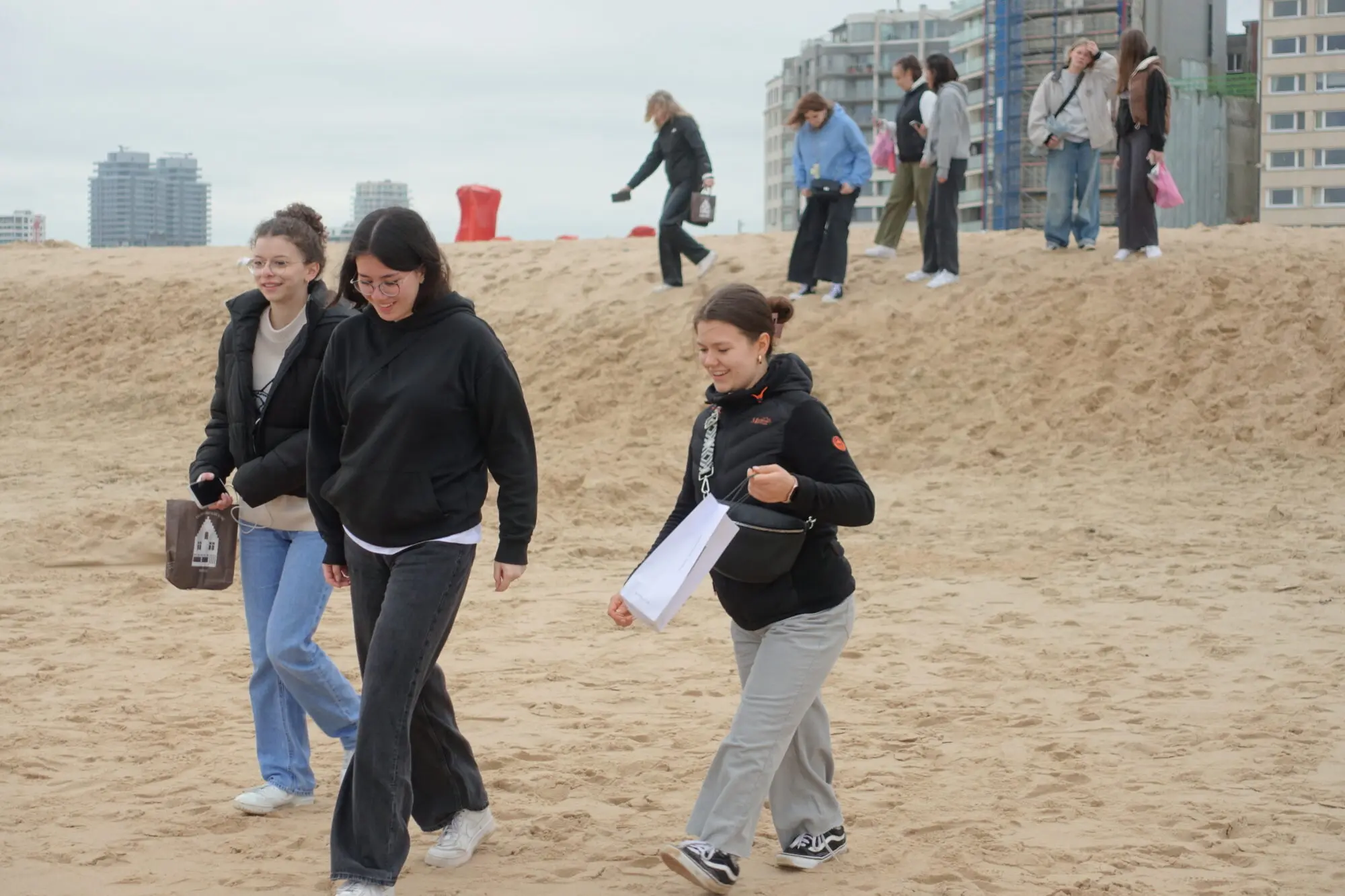 Jugendliche spazieren an einem Sandstrand, im Hintergrund Gebäude einer Küstenstadt