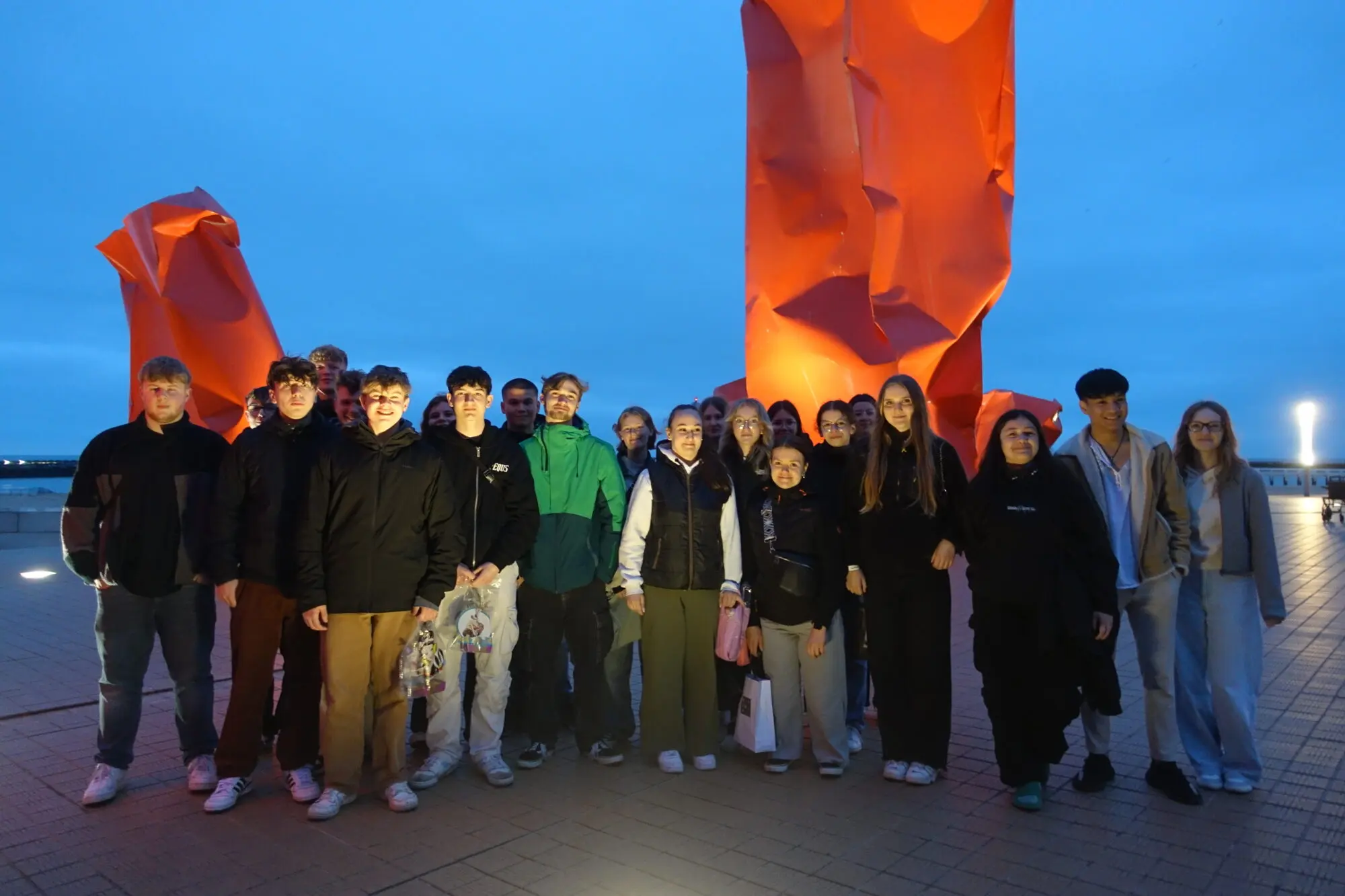 Schülergruppe posiert in der Abenddämmerung vor einer großen roten Skulptur
