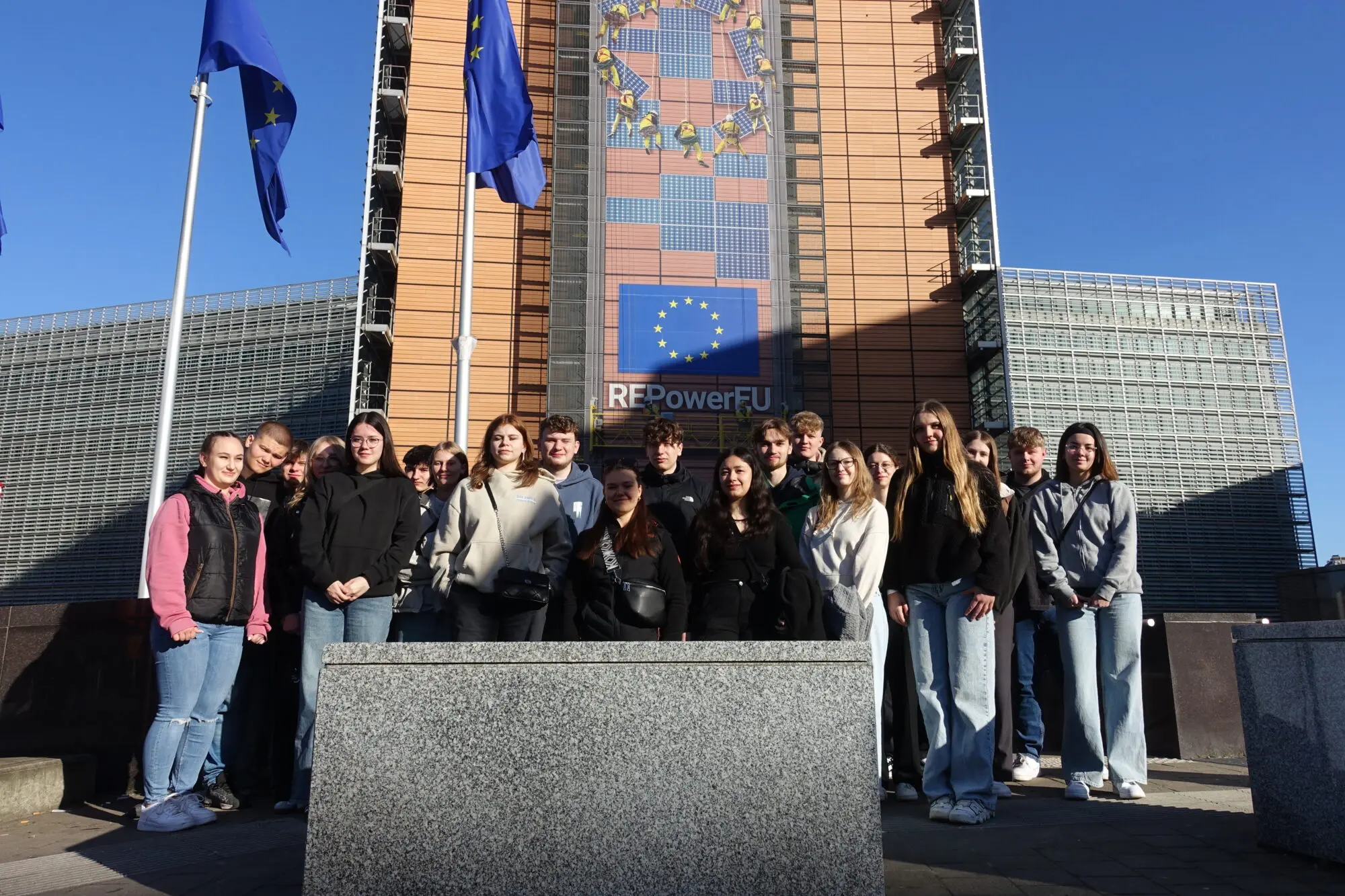 Schülergruppe posiert vor einem EU-Gebäude in Brüssel mit Europafahnen bei blauem Himmel
