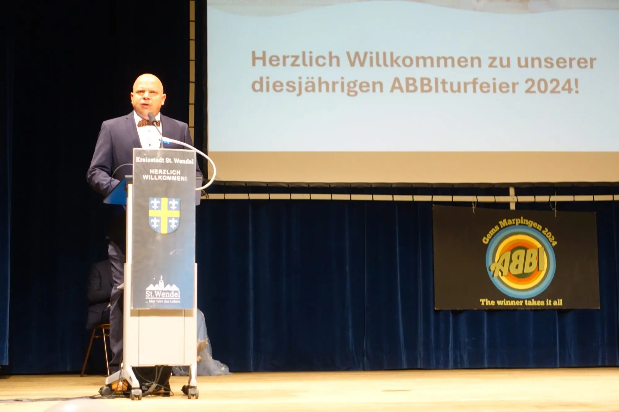 Redner am Pult auf einer Bühne, Leinwand: „Herzlich Willkommen zu unserer diesjährigen Abiturfeier 2024!“