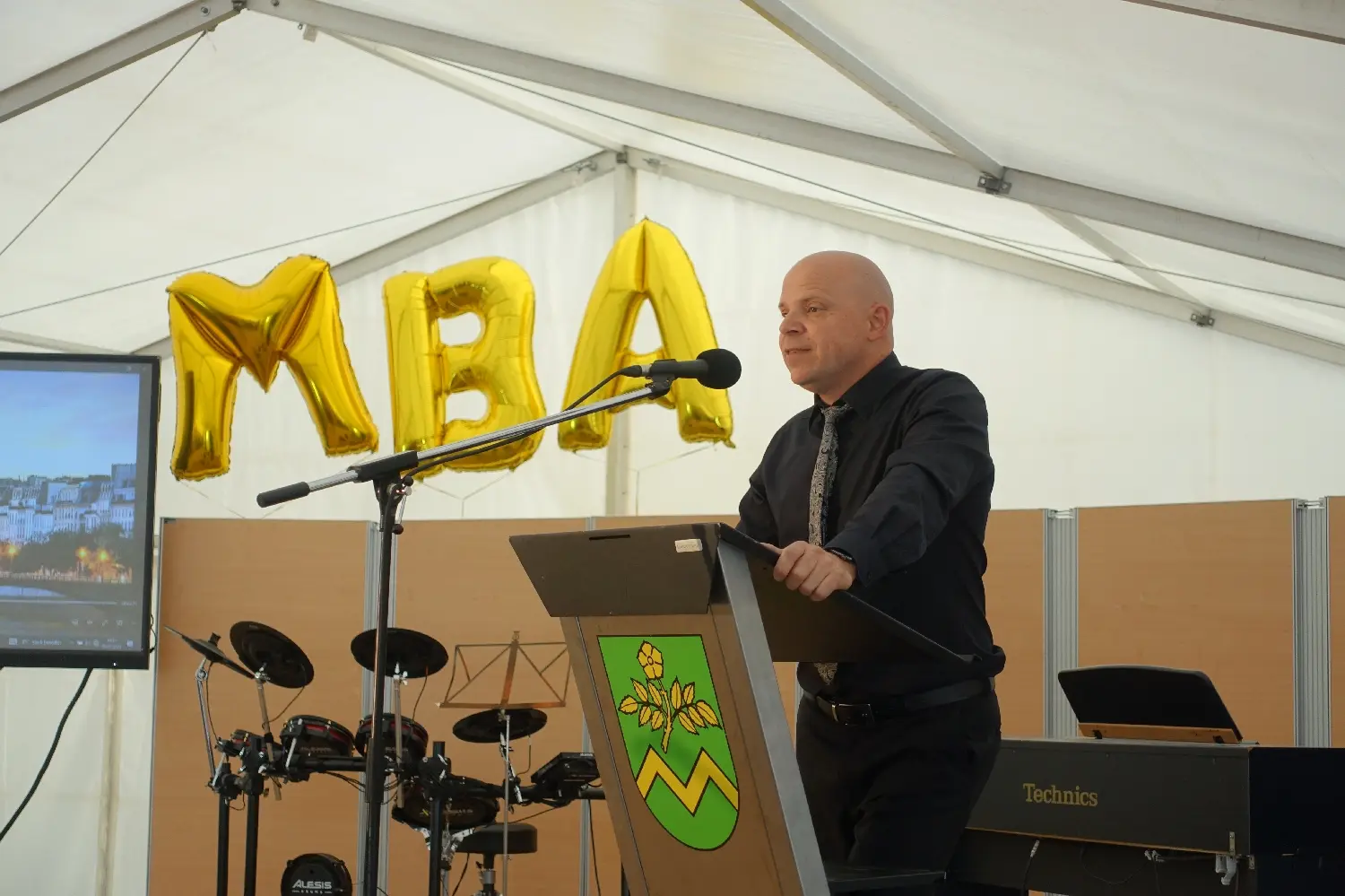 Redner am Pult im Festzelt, dahinter goldene Buchstaben „MBA“ und ein Schlagzeug