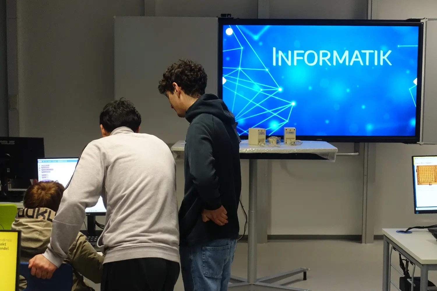 Kinder und Jugendliche an Computern vor einem großen Bildschirm mit der Aufschrift „Informatik“