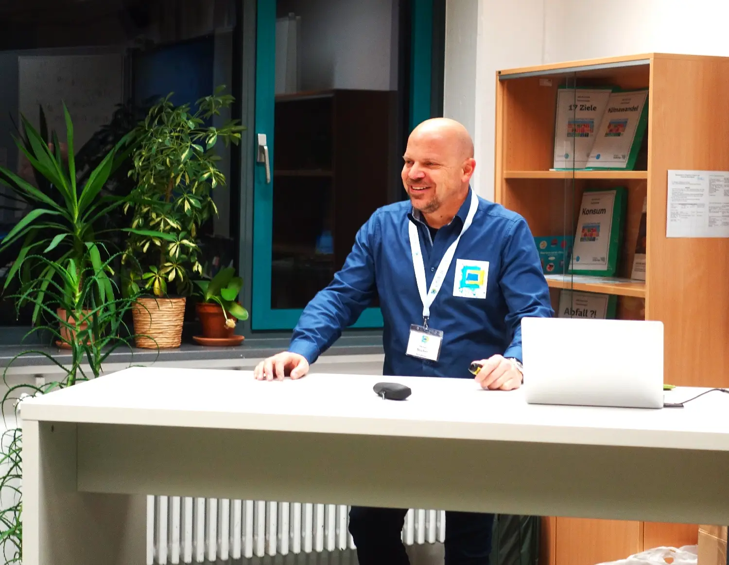Mann mit Namensschild sitzt an einem Schreibtisch mit Laptop und lächelt