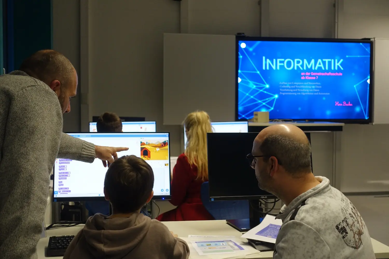 Kinder und Erwachsene an Computern im Informatikraum, auf dem Bildschirm steht „Informatik“