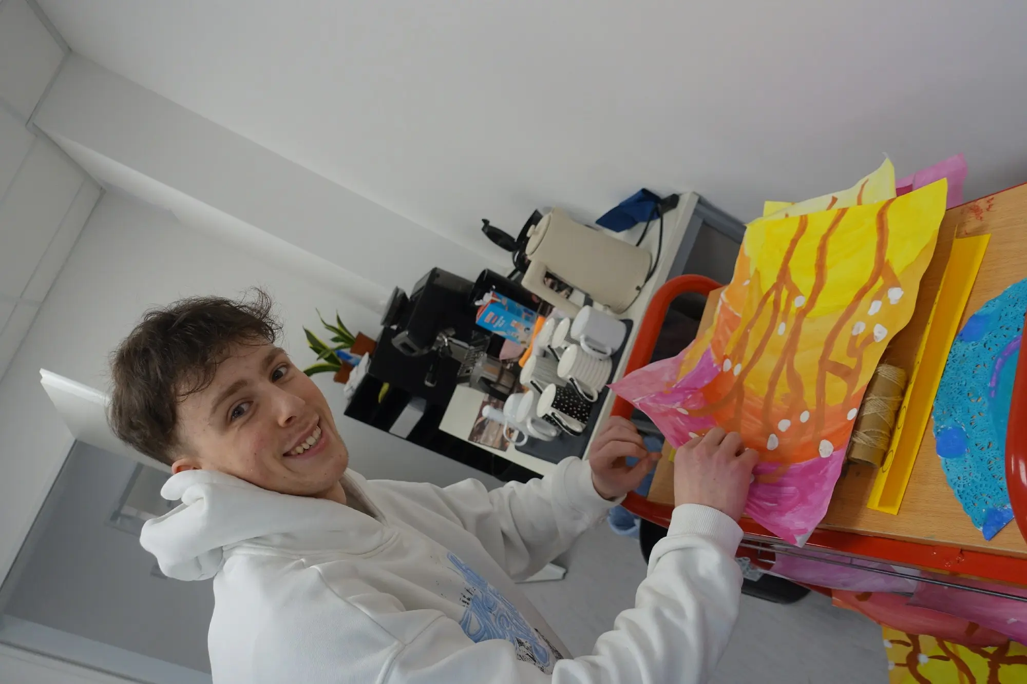Jugendlicher räumt bunte Bastelarbeiten aus Papier in ein Regal in einem Gruppenraum