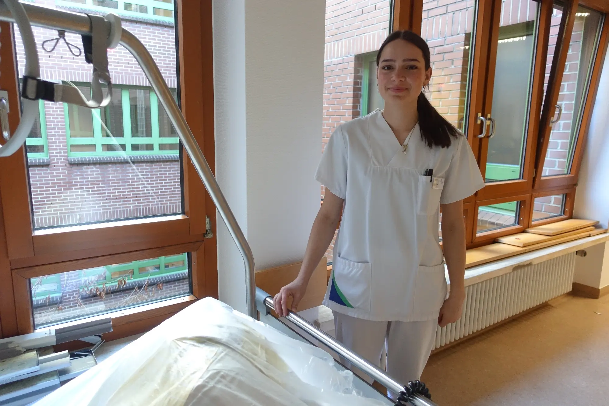 Junge Frau in weißer Pflegekleidung steht neben einem Pflegebett in einem Zimmer