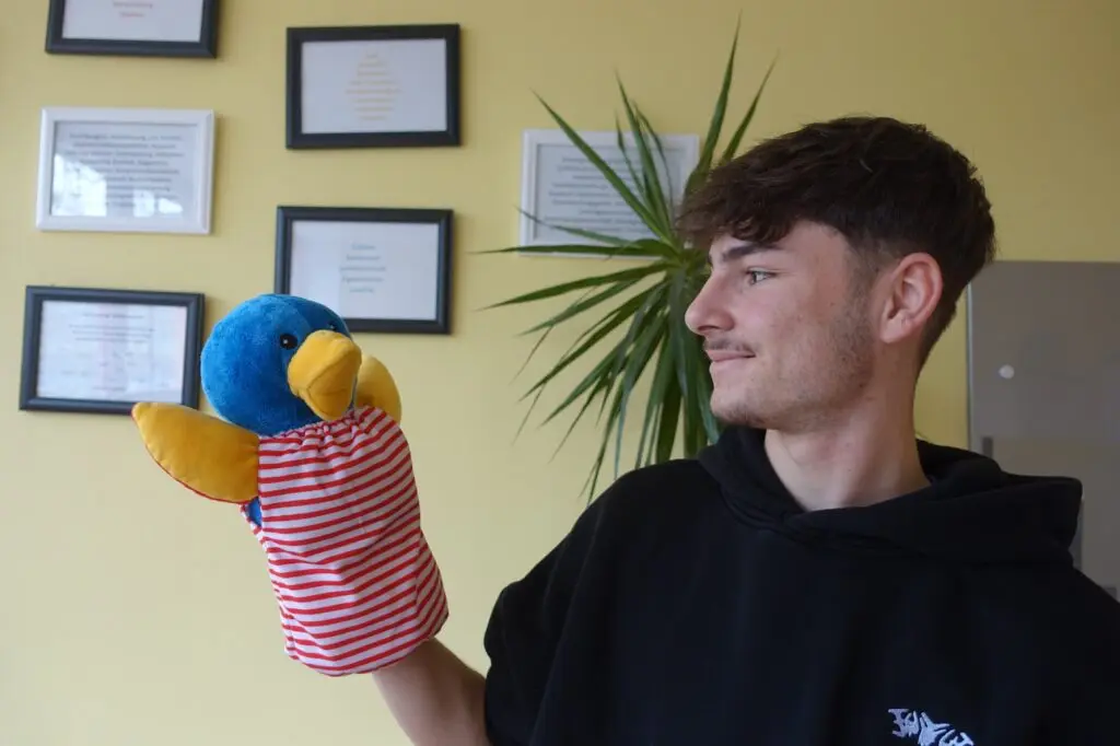 Jugendlicher hält lachend eine bunte Vogel-Handpuppe in einem Raum mit Bildern an der Wand