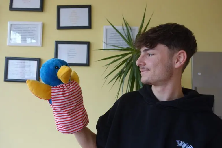 Jugendlicher hält lachend eine bunte Vogel-Handpuppe in einem Raum mit Bildern an der Wand