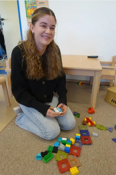 Junge Frau sitzt lächelnd auf dem Boden eines Spielraums neben bunten Bauklötzen