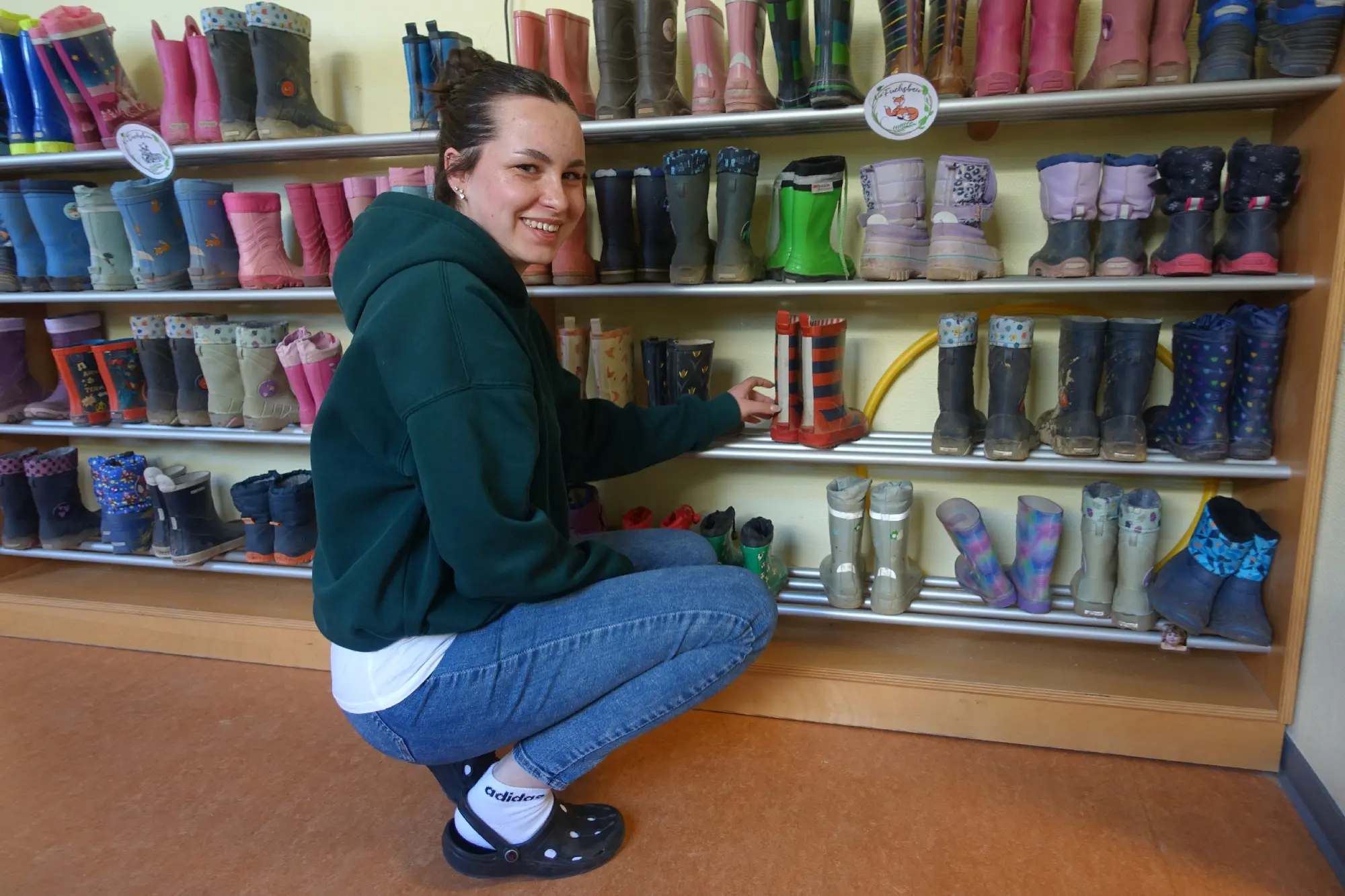Junge Frau hockt lächelnd vor einem langen Regal voller bunter Kindergummistiefel