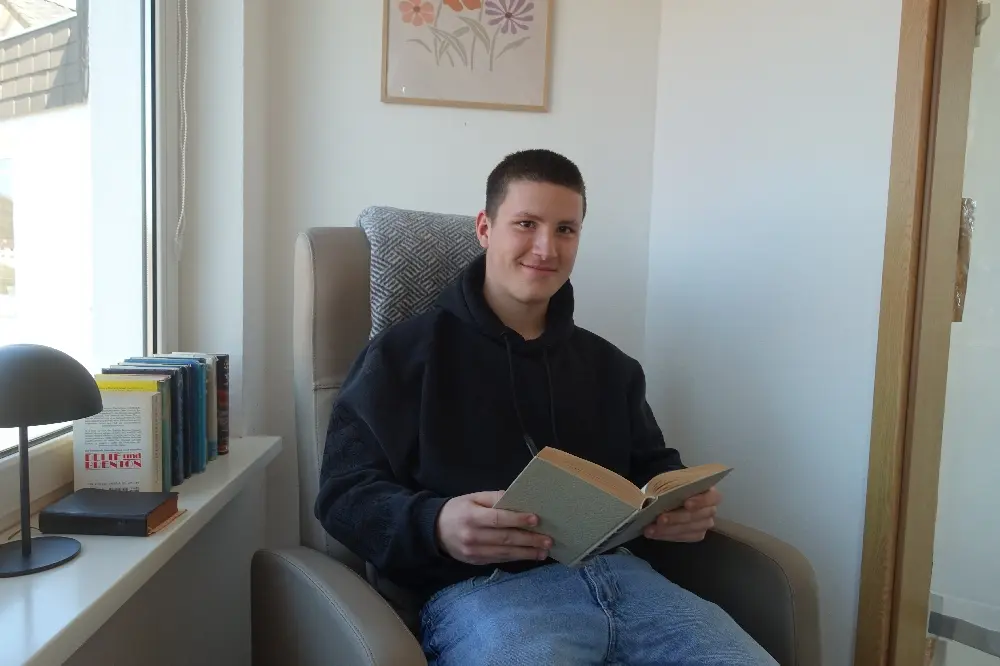 Jugendlicher sitzt in einem Sessel am Fenster und liest in einem Buch