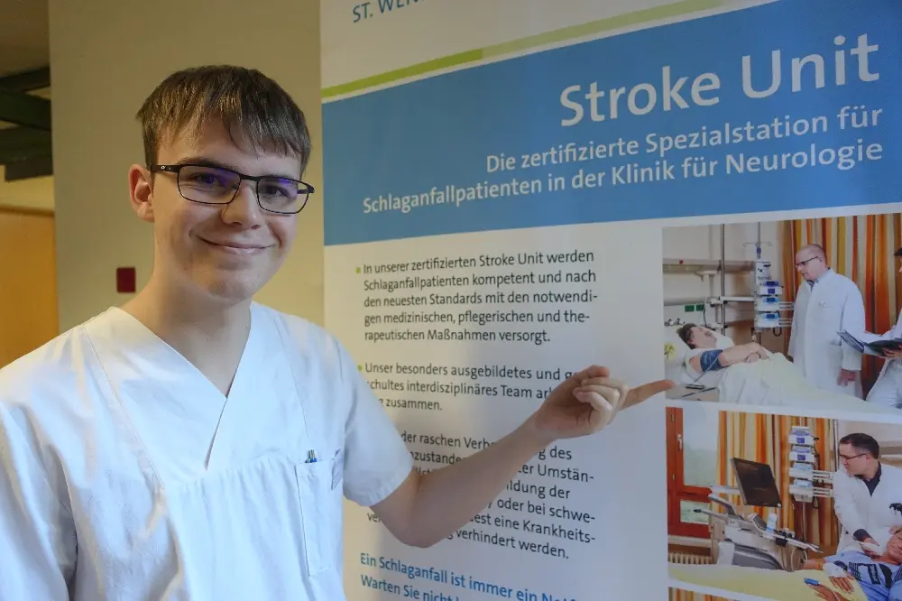 Junger Mann in weißer Kleidung zeigt auf eine Infotafel „Stroke Unit“