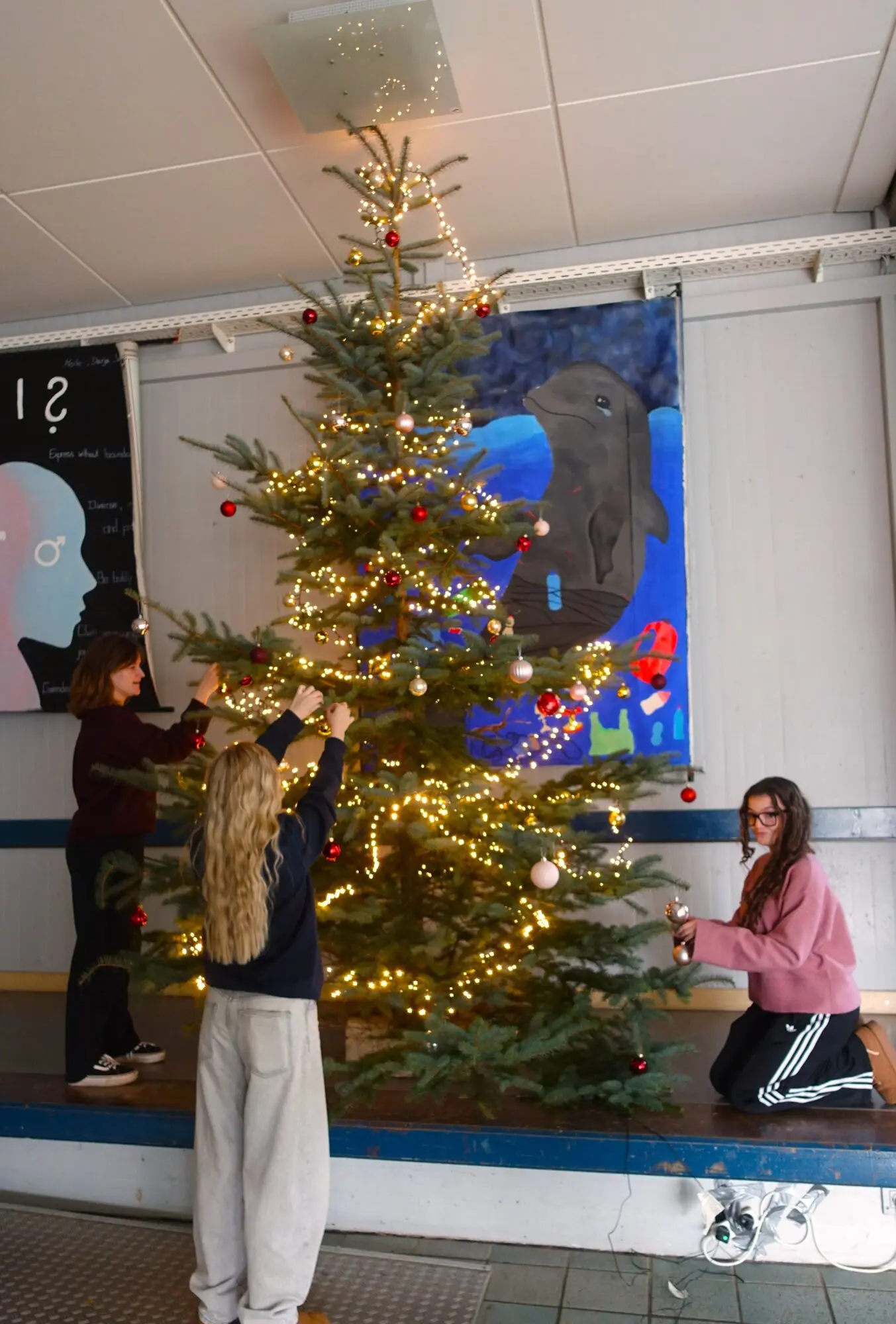 Beleuchteter Weihnachtsbaum im Schulfoyer, Schülerinnen hängen Schmuck auf