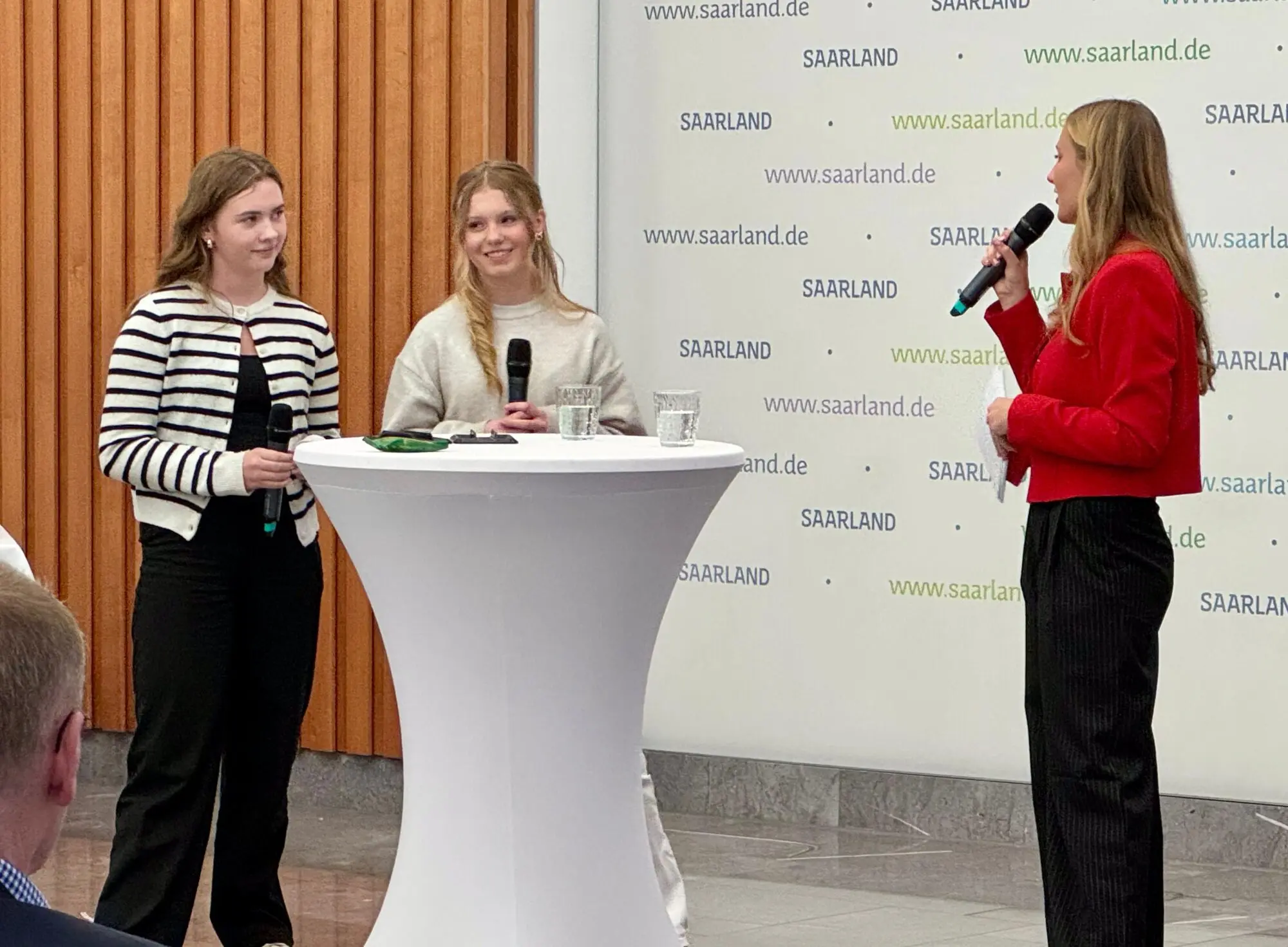 Zwei Schülerinnen an einem Stehtisch im Gespräch mit einer Moderatorin mit Mikrofon
