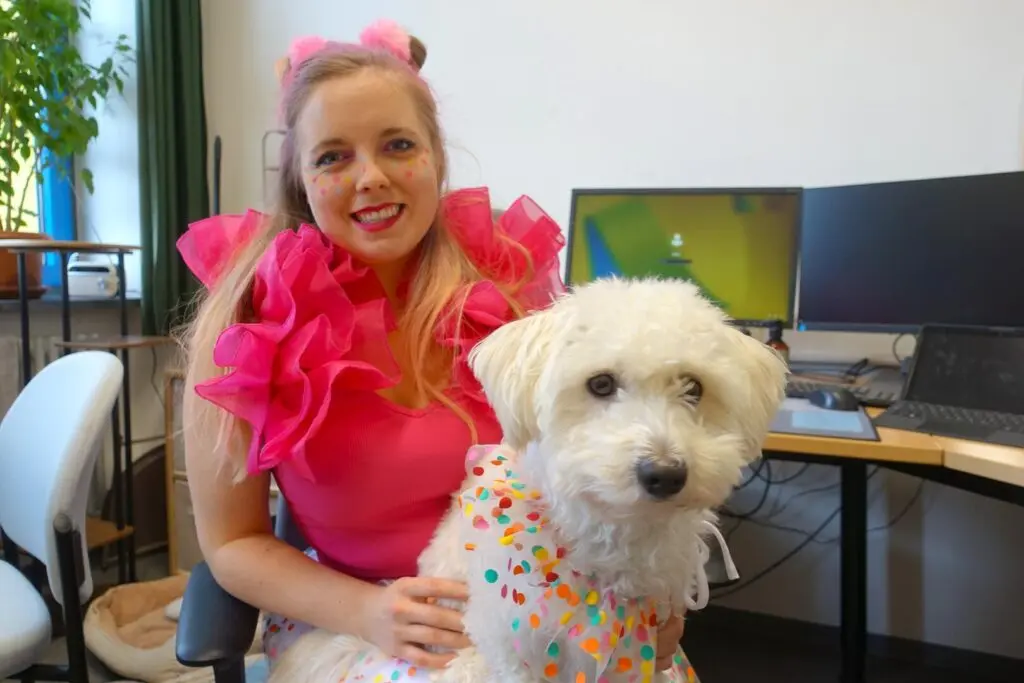 Frau in pinkem Rüschenoberteil sitzt mit einem weißen Hund mit buntem Halstuch in einem Büro