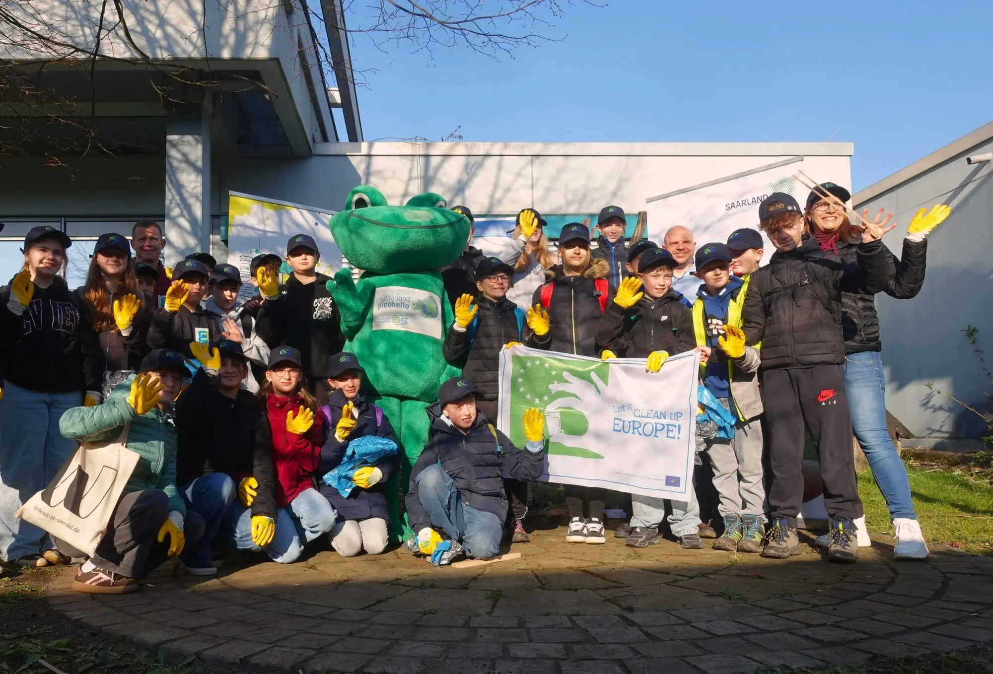 Große Schülergruppe mit grünem Maskottchen und Banner bei einer Müllsammelaktion