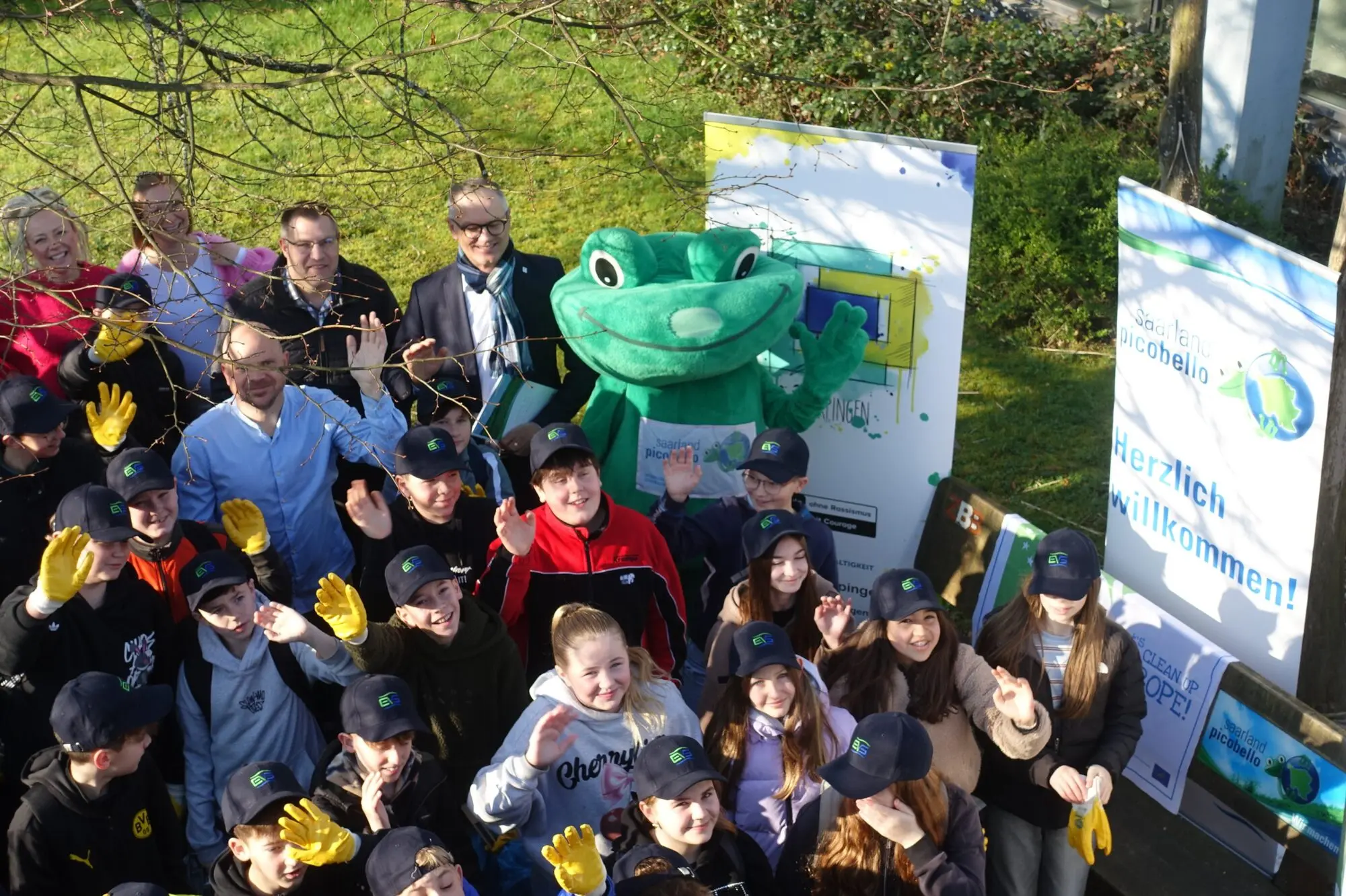 Große Schülergruppe posiert gemeinsam mit einem Frosch-Maskottchen vor Werbebannern im Freien