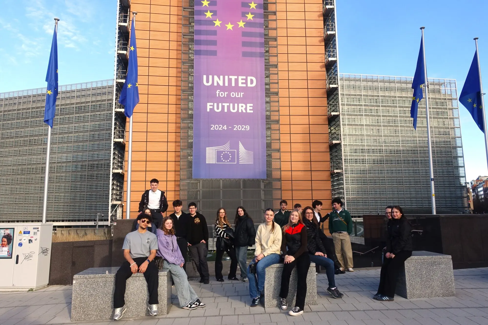 Schülergruppe sitzt und steht vor einem EU-Gebäude mit Europaflaggen und Banner „United for our future“