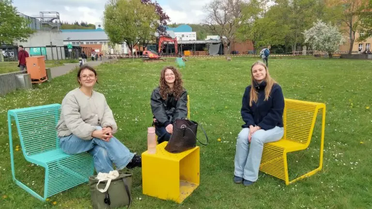 Drei Jugendliche sitzen auf bunten Sitzmöbeln auf einer Wiese vor dem Schulgelände
