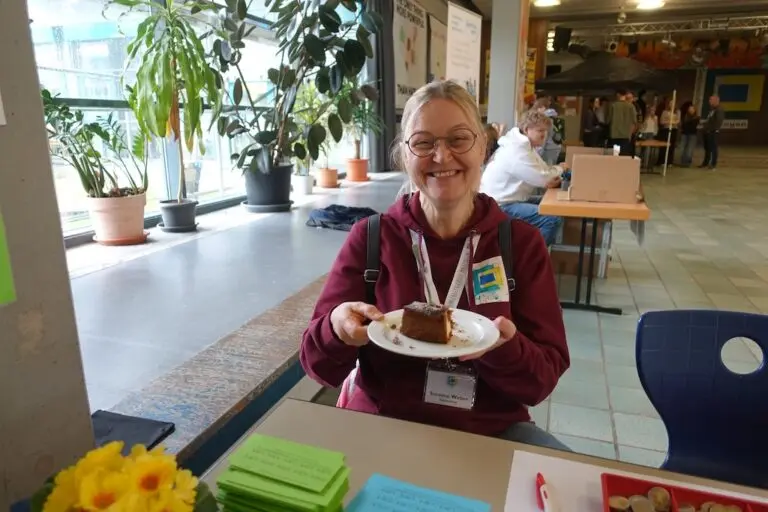 Frau mit Namensschild sitzt lächelnd mit einem Teller Kuchen an einem Tisch
