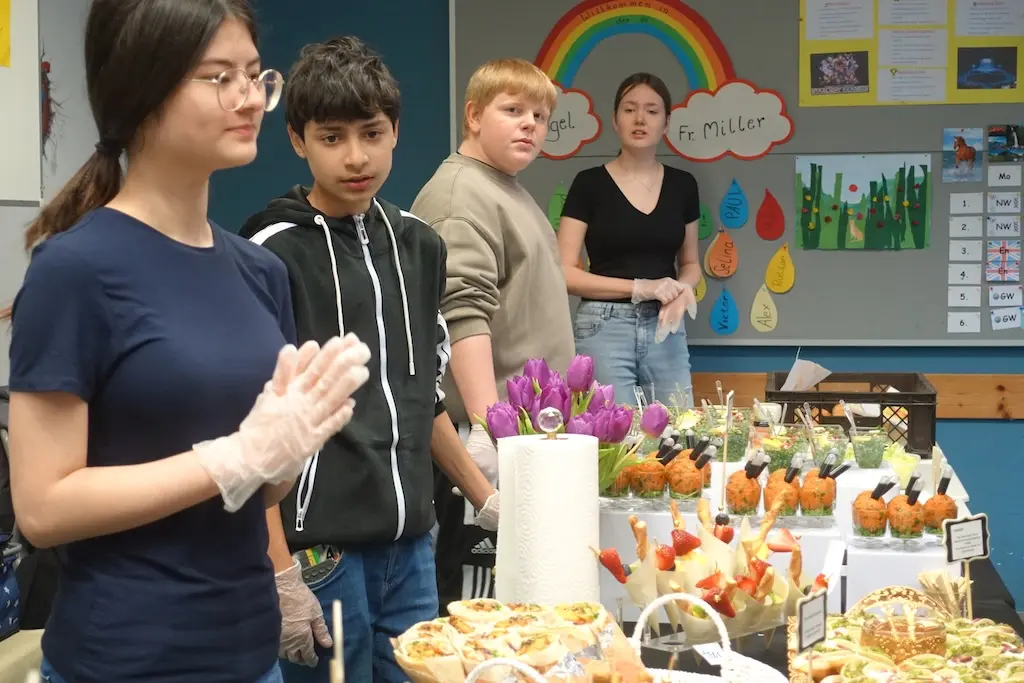 Jugendliche stehen an einem Buffet mit Snacks und Blumen im Schulraum