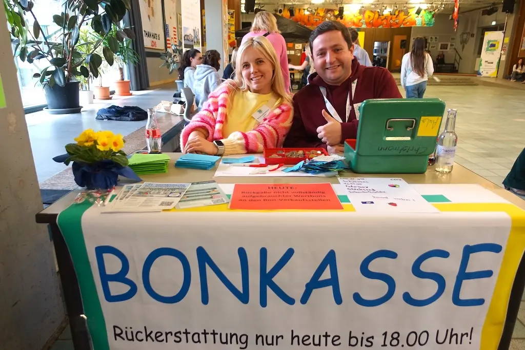 Zwei Frauen sitzen lachend hinter dem Tisch der Bonkasse mit Banner