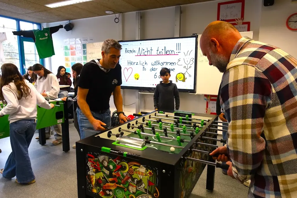 Erwachsene spielen Tischkicker, im Hintergrund Bildschirm und Besucher