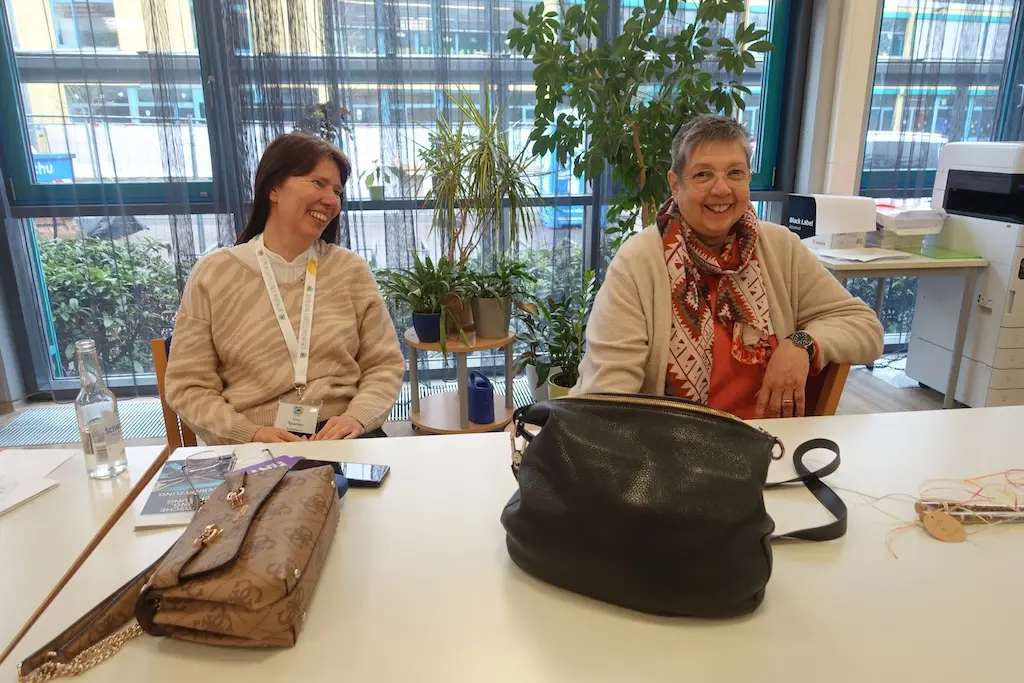 Zwei Frauen mit Namensbändern sitzen an einem Tisch mit Taschen