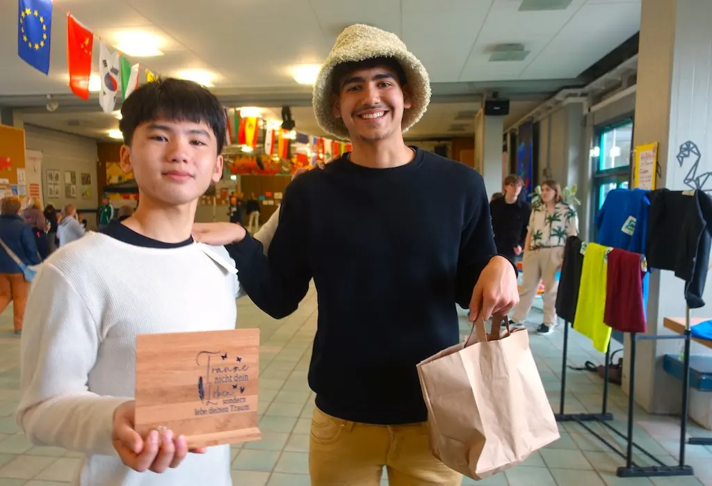 Zwei Jugendliche zeigen ein Holzschild und eine Tüte in der Pausenhalle