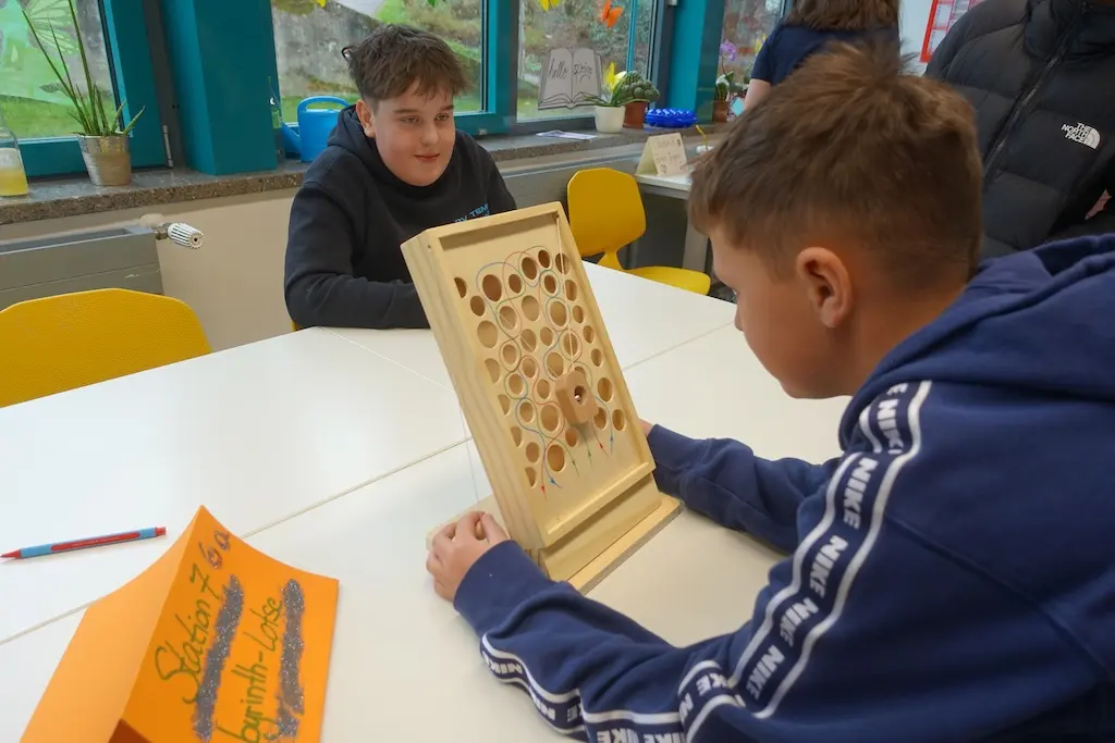 Schüler sitzt an einem Tisch und hält ein Holzspiel mit Kugeln hoch