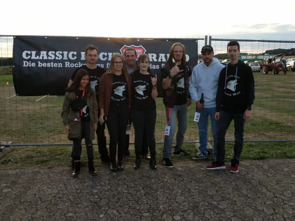 Gruppe Jugendlicher und Erwachsener vor einem Banner einer Rockveranstaltung auf einem Festivalgelände
