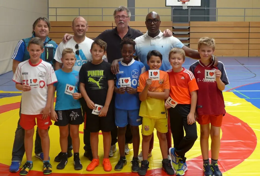 Kinder in bunten Trikots stehen mit Erwachsenen als Gruppe auf einer Ringermatte in der Sporthalle