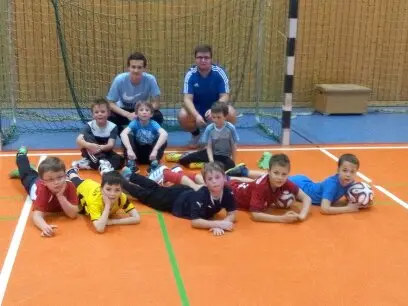 Kinder in Sportkleidung liegen und sitzen gruppiert auf dem Boden einer Sporthalle