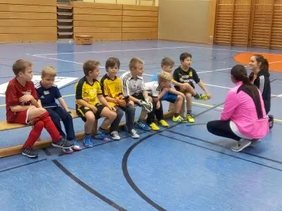 Kinder sitzen auf einer Bank in der Sporthalle, davor hockt eine junge Trainerin