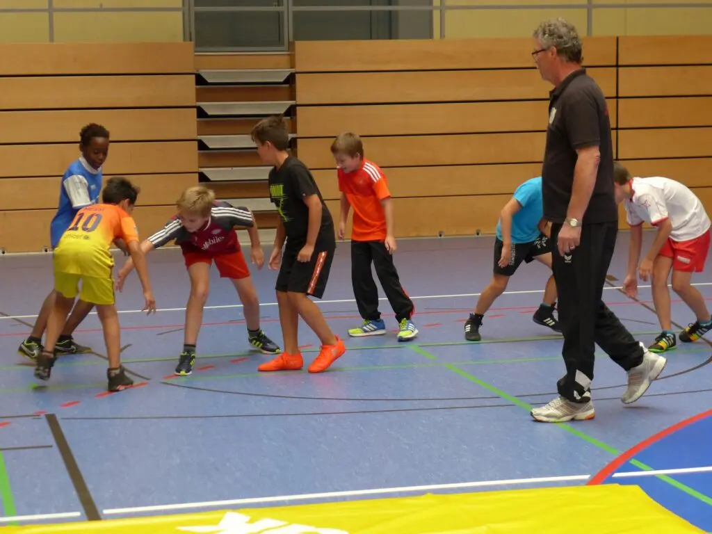 Kinder machen Aufwärmübungen auf dem Hallenboden, ein Trainer leitet an