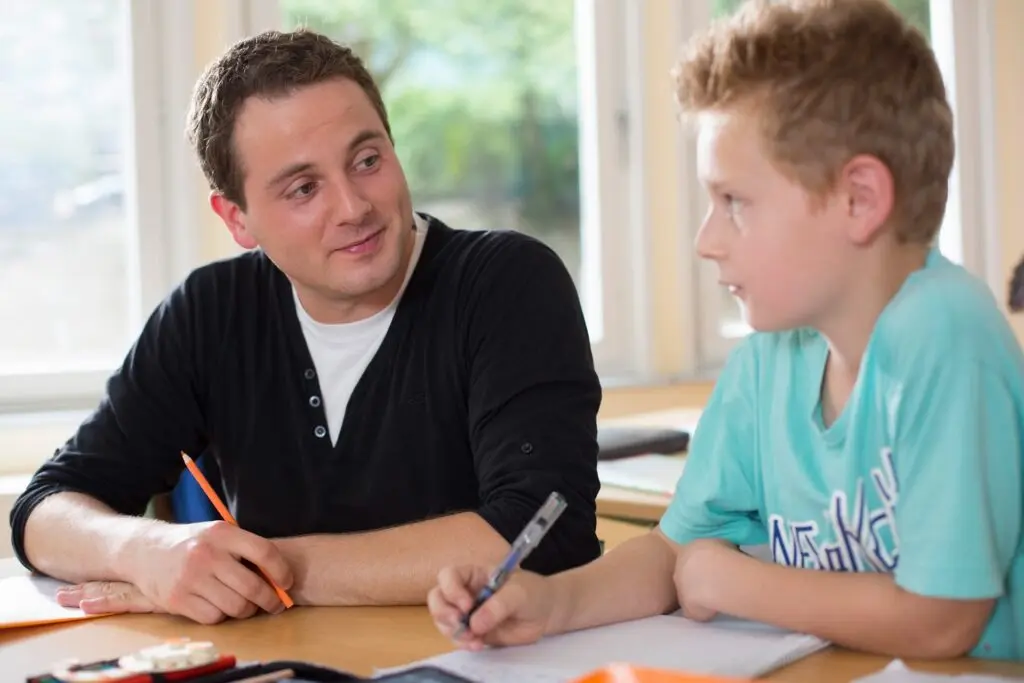 Junger Mann und Schüler sitzen mit Stiften und Heften am Tisch und sprechen über eine Aufgabe
