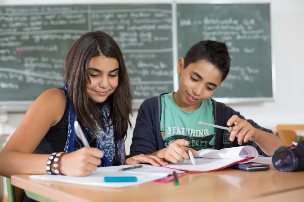 Schülerin und Schüler schreiben gemeinsam in Hefte an einem Tisch vor einer Tafel