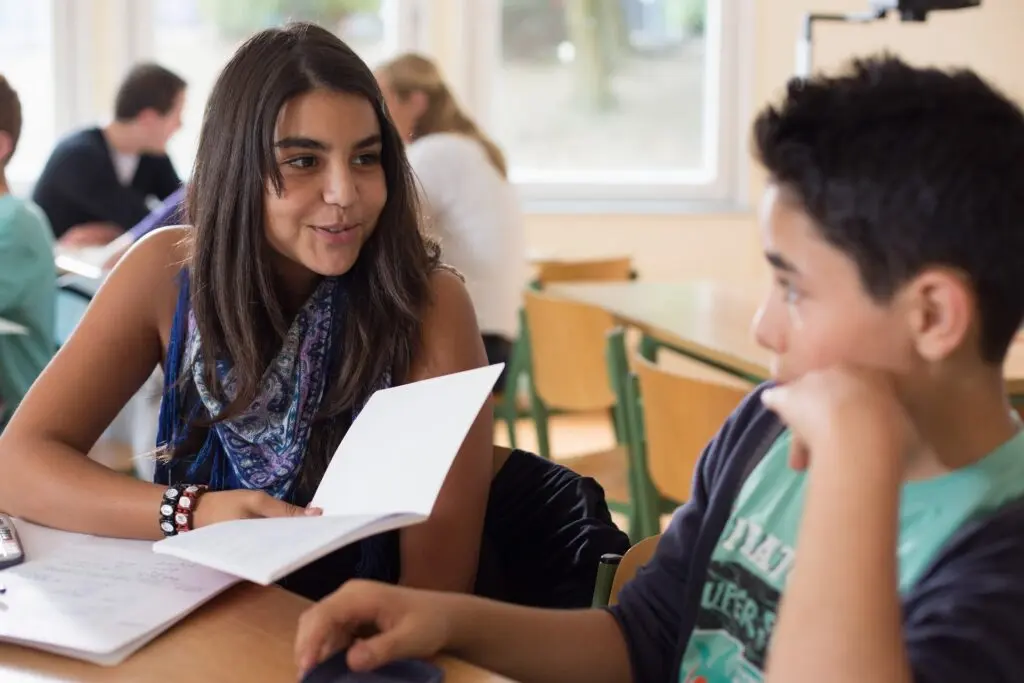 Schülerin und Schüler sitzen mit Heften an einem Tisch im Klassenraum und unterhalten sich beim Lernen