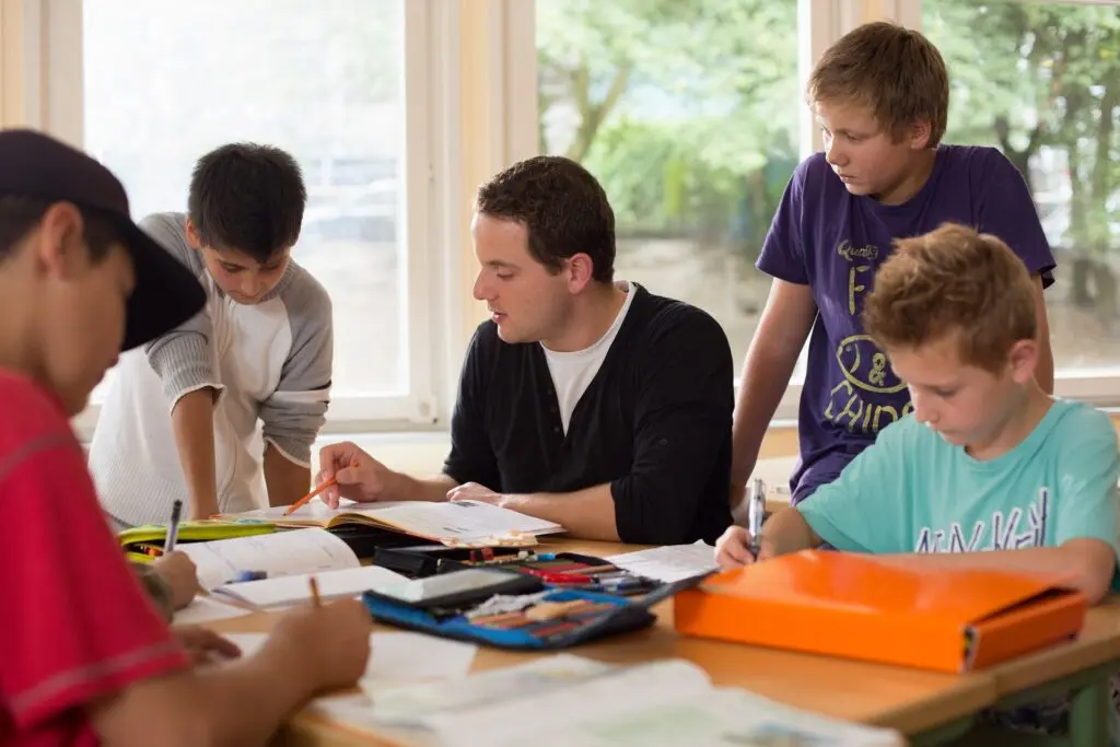 Schüler arbeiten an einem Tisch mit Heften, ein Erwachsener hilft bei der Lernförderung