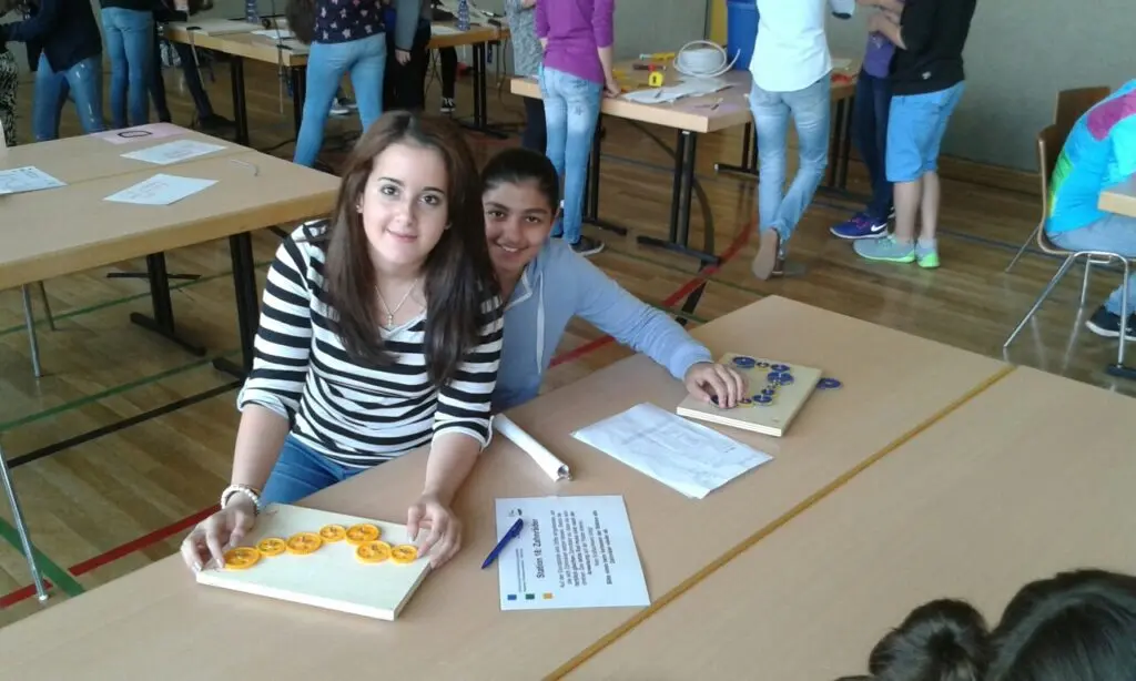 Zwei Schülerinnen sitzen lächelnd an einem Tisch mit Arbeitsblättern und einer bemalten Platte
