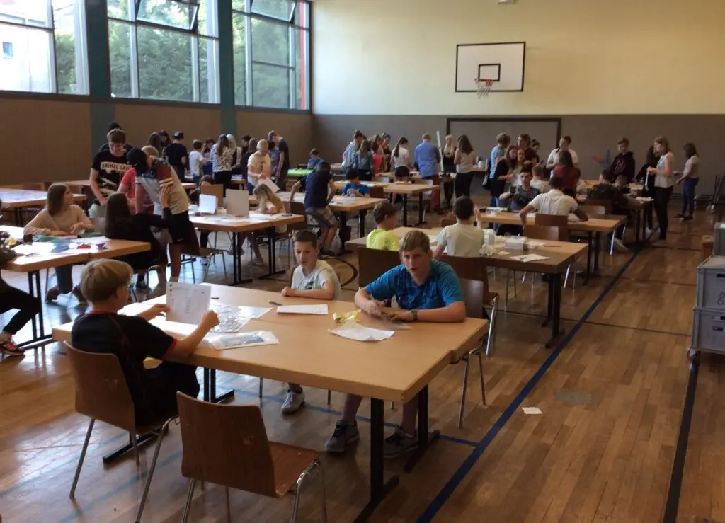 Viele Schülerinnen und Schüler sitzen an Tischreihen in einer Sporthalle und bearbeiten praktische Aufgaben