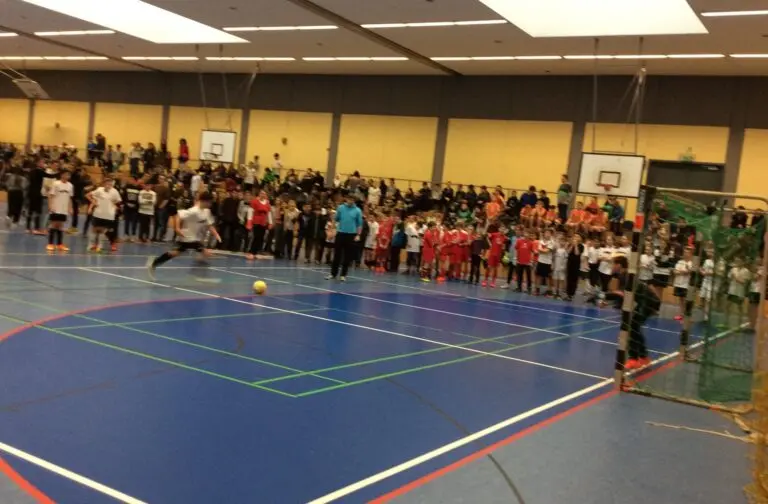 Fußballspiel in einer Sporthalle, auf der Tribüne sitzen zahlreiche Zuschauerinnen und Zuschauer