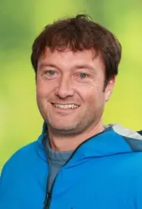 Porträt Nicolas Andrée, lächelnd in blauer Jacke vor grünem Hintergrund
