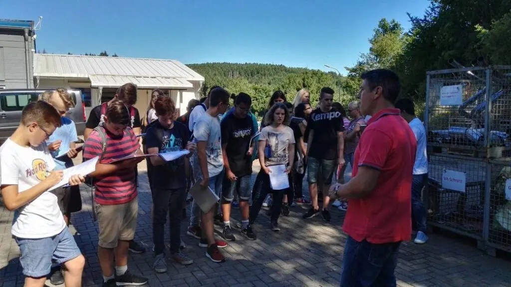 Schülergruppe mit Arbeitsblättern hört im Freien einem Mann vor einem Raiffeisenmarkt zu