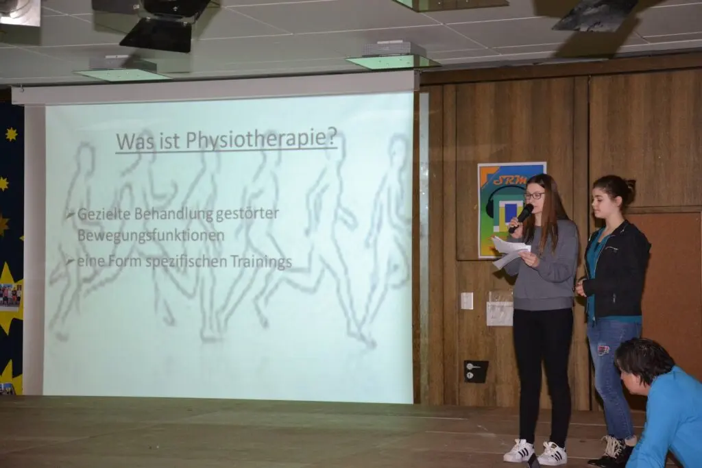 Zwei Schülerinnen präsentieren auf der Bühne vor einer Leinwand mit Folie zum Physiotherapeuten