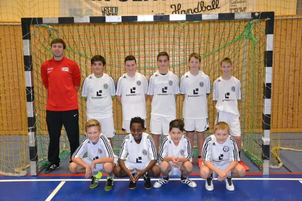 Kindermannschaft in weißen Trikots mit Trainer in Rot vor einem Handballtor in der Sporthalle