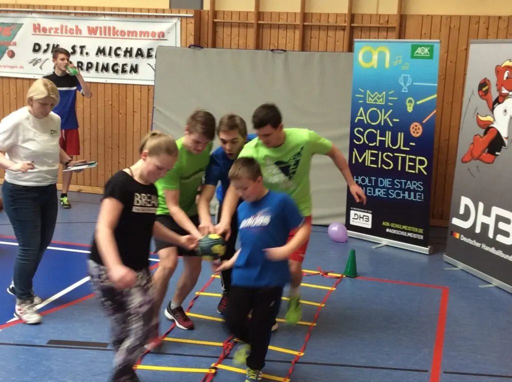 Schülergruppe hält gemeinsam einen Handball und läuft durch einen Parcours in der Halle, dahinter Banner „AOK-Schulmeister“ und DHB