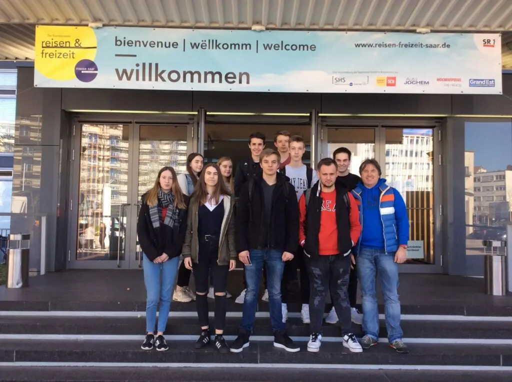 Schülergruppe steht mit Begleitpersonen auf der Treppe vor einem Gebäudeeingang mit Banner „willkommen“
