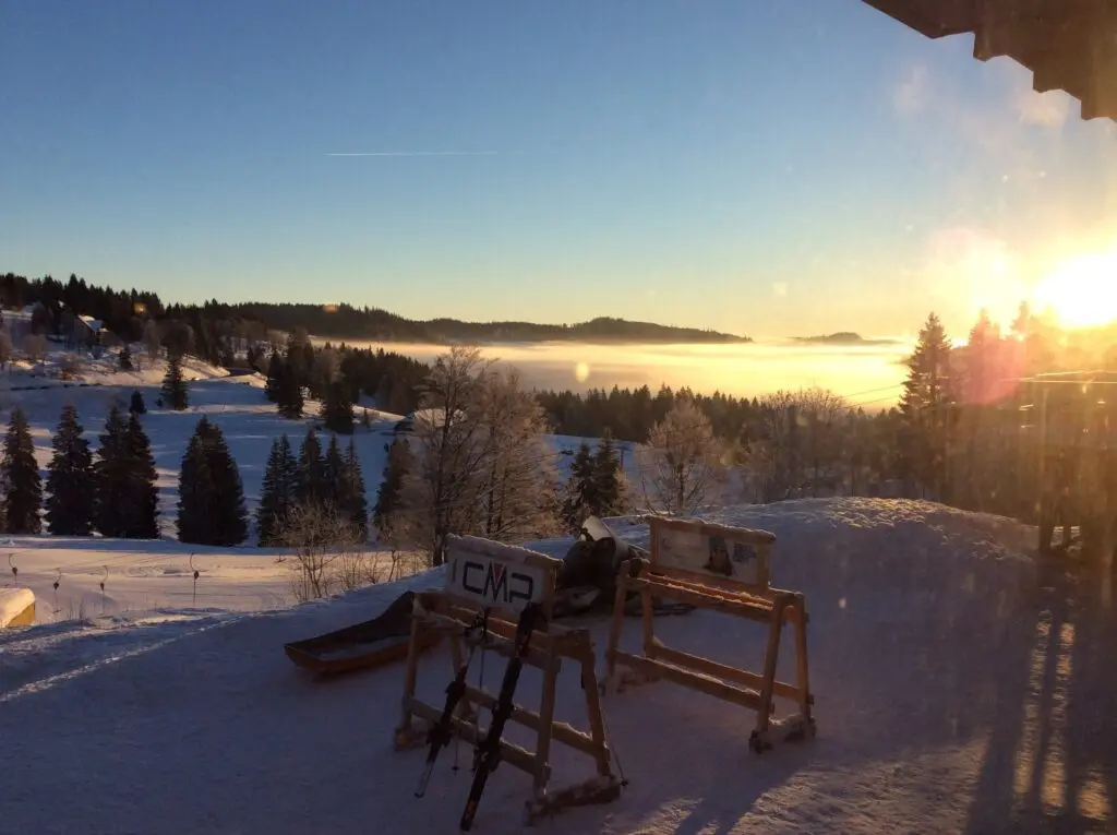 Sonnenaufgang über verschneiter Berglandschaft mit Nebel im Tal, vorne Holzgeländer und Schnee